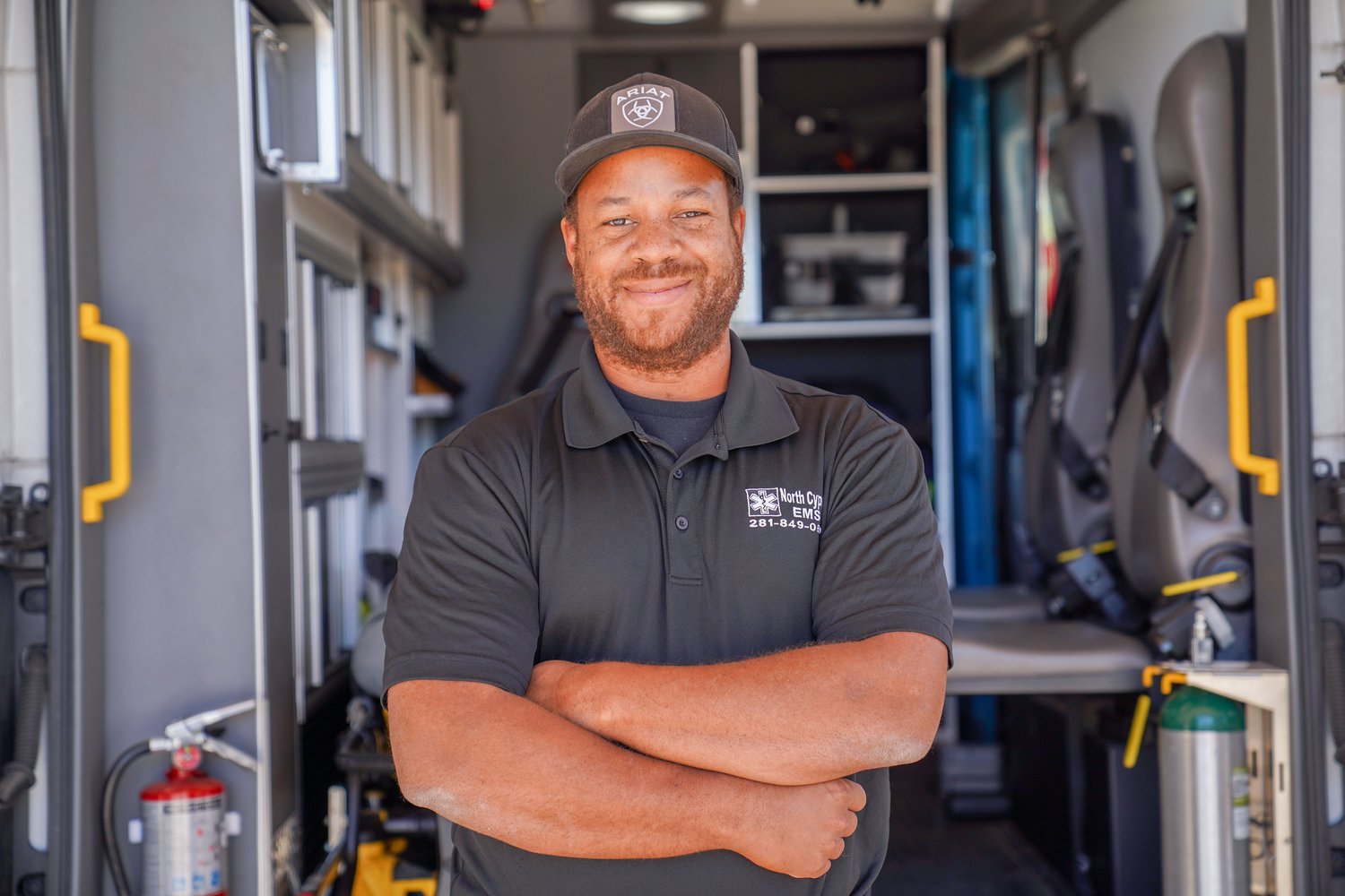 Man with arms crossed wearing a hat smiling in front of an opened ambulance